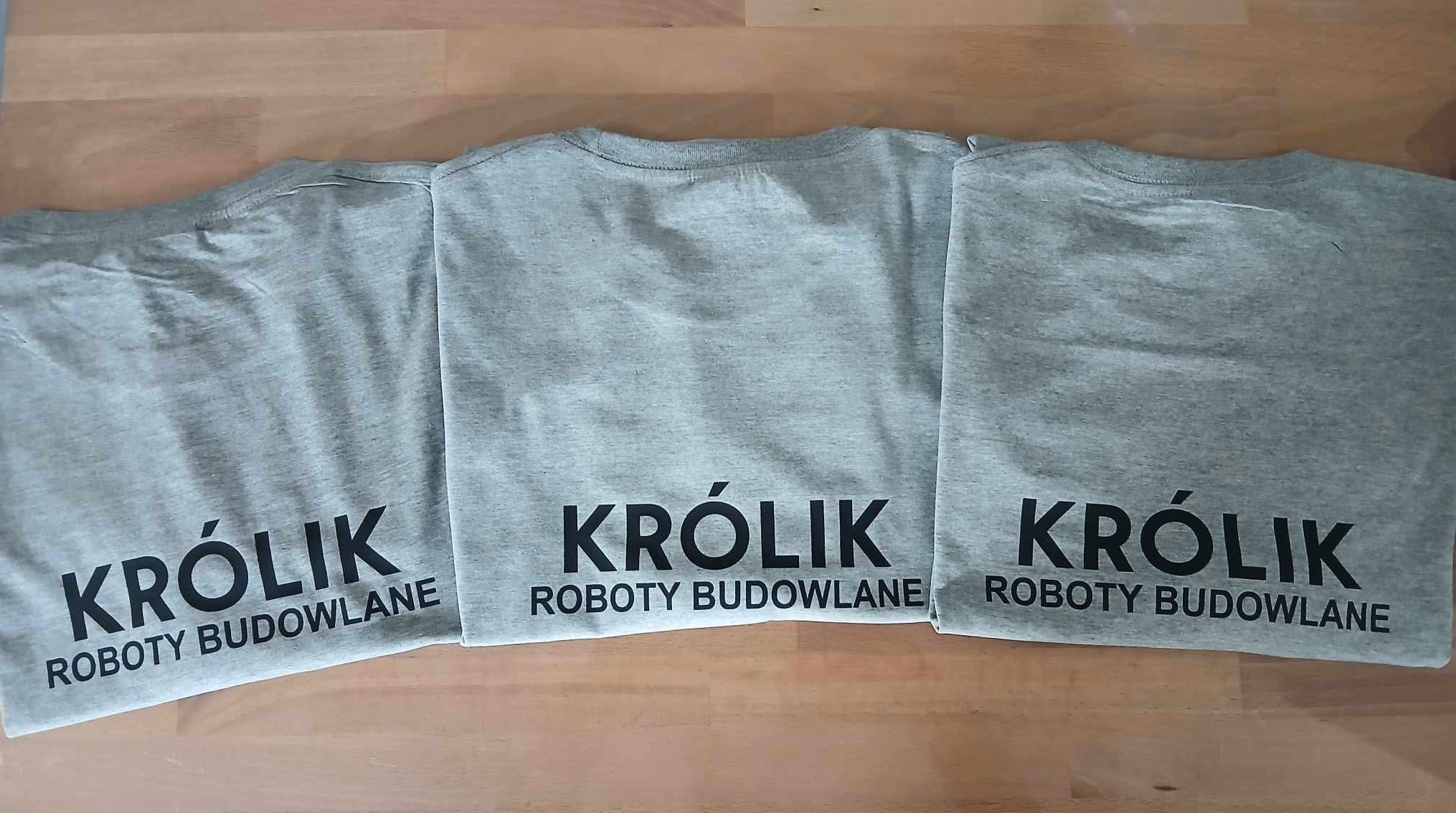 Trzy szare koszulki z czarnym nadrukiem 'KRÓLIK ROBOTY BUDOWLANE' leżące obok siebie na drewnianym blacie.