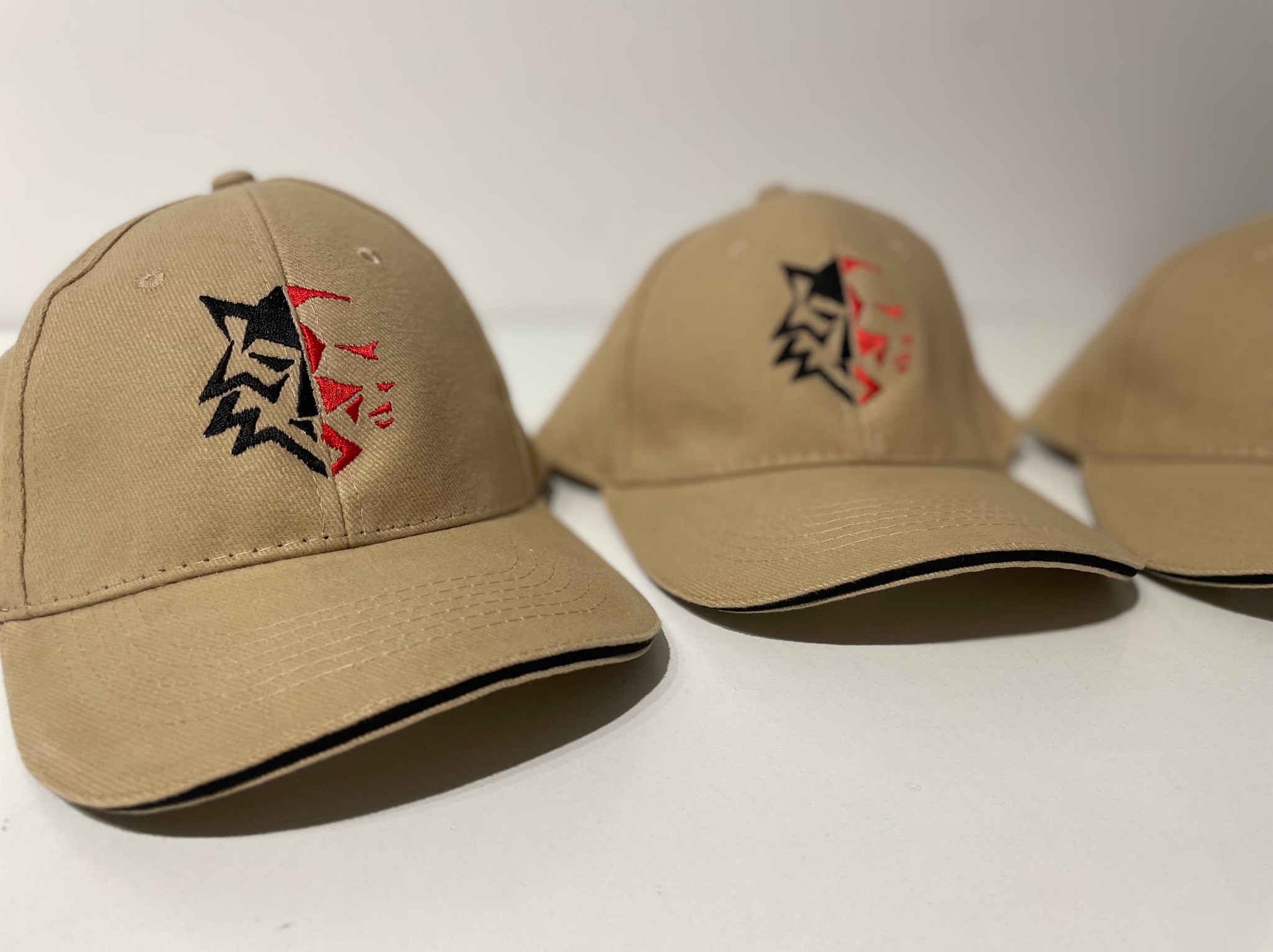Trzy beżowe czapki z daszkiem z wyhaftowanym czarno-czerwonym logo na froncie, leżące na białym stole.