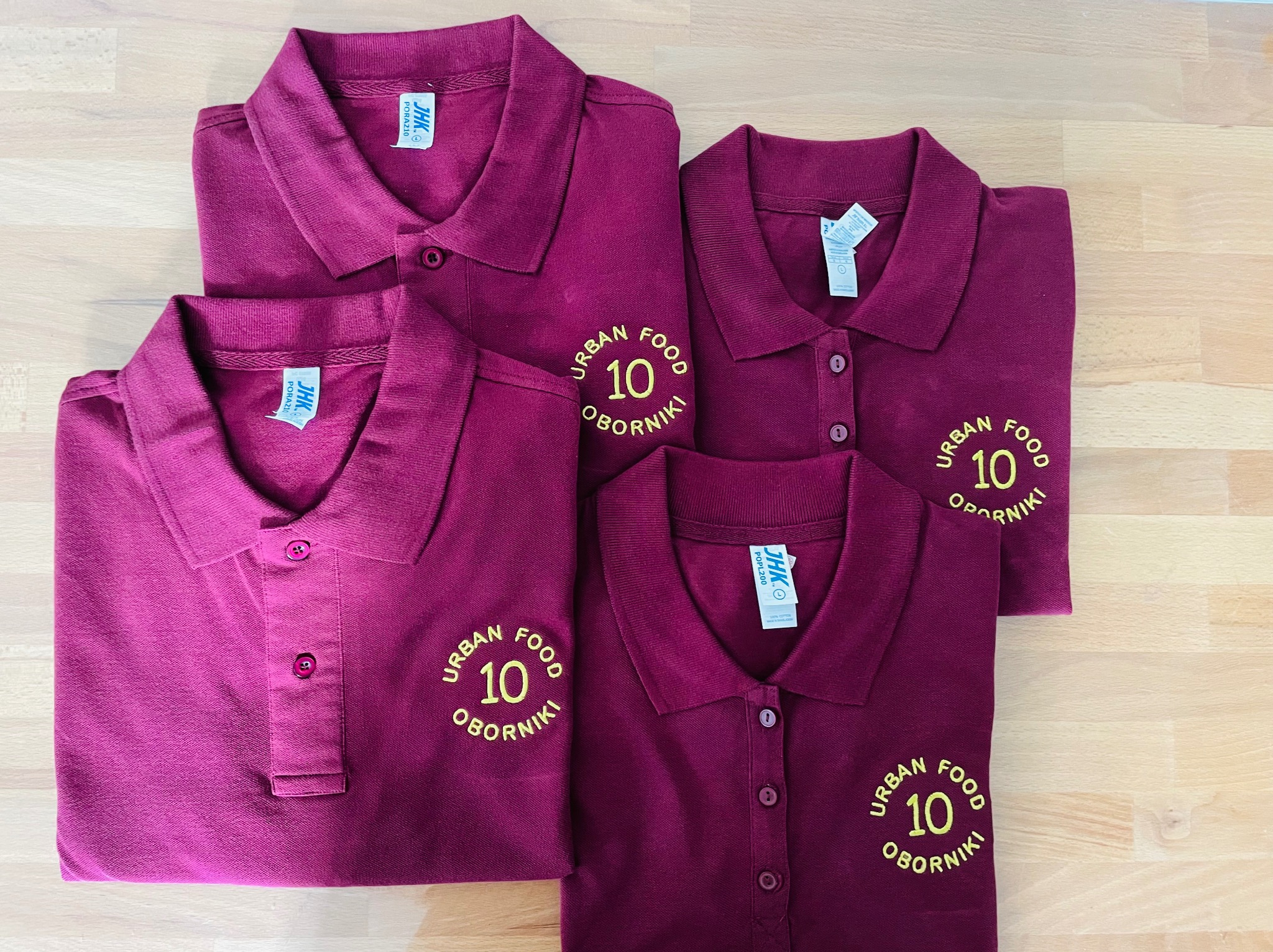 Cztery bordowe koszulki polo z haftowanym logo 'URBAN FOOD 10 OBORNIKI' ułożone na drewnianym blacie.