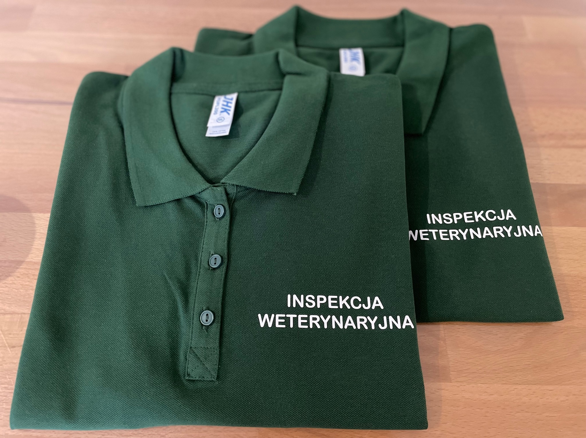 Dwa zielone polo z białym napisem 'Inspekcja Weterynaryjna' leżące na drewnianym blacie, widoczne guziki i metka.