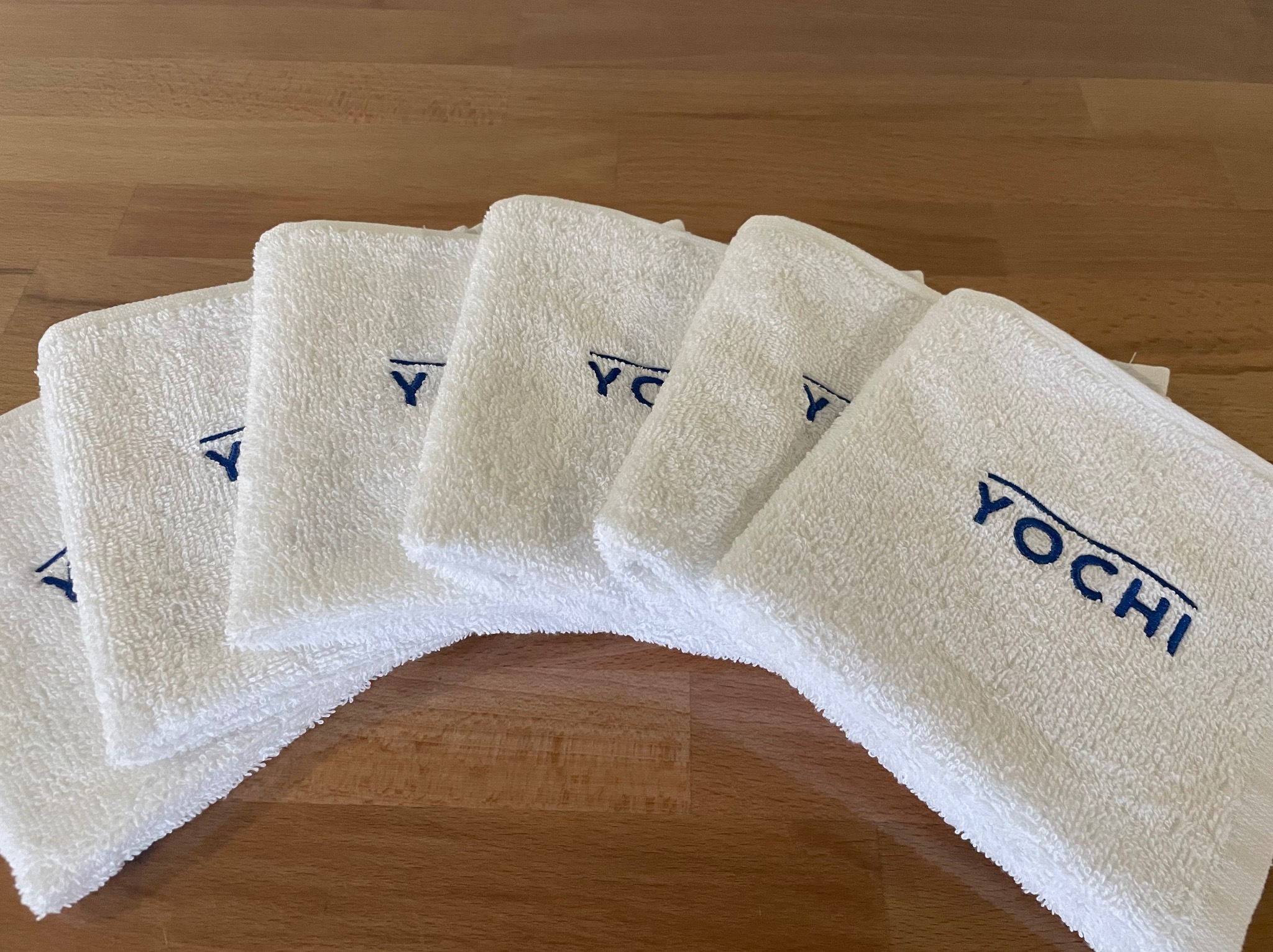 Ułożone rzędem białe ręczniki frotte z wyhaftowanym granatowym napisem 'YOCHI' na drewnianym blacie.