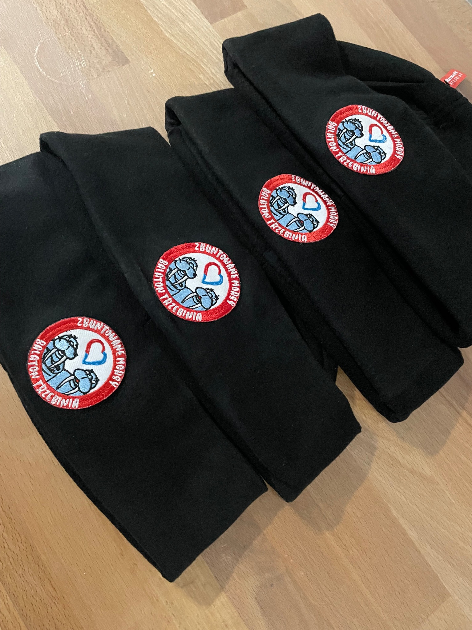 Czarne bluzy polarowe ułożone na drewnianym podłożu, ozdobione okrągłymi naszywkami z logo 'Zbuntowane Morsy Balaton Trzebinia'.