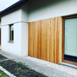 Elewacja domu z jasnego tynku z drewnianą okładziną z pionowych desek, przylegająca do okna z żaluzjami, z chodnikiem z kostki brukowej i rabatką.