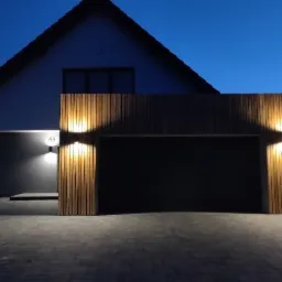 Nowoczesna elewacja domu z garażem obłożona pionowymi panelami drewnianymi, oświetlona dwoma reflektorami ściennymi, widoczne numer domu '63' i kostka brukowa przed garażem, sfotografowane...