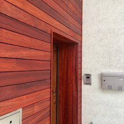 RedWood - Ciemnoczerwona drewniana elewacja domu z poziomymi deskami, przylegająca do białej ściany z domofonem i skrzynką na listy, obok drewniane obramowanie drzwi.