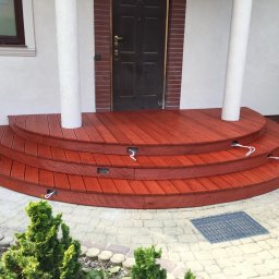 RedWood - Trzy poziomy schodów wejściowych z czerwonego drewna prowadzące do ciemnych drzwi wejściowych, otoczone białymi kolumnami, z widocznymi przewodami oświetleniowymi i kostką brukową na zewnątrz.