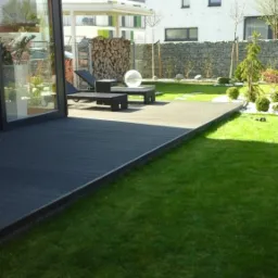 Nowoczesny taras z kompozytu w kolorze grafitowym, przylegający do budynku z dużym przeszkleniem, otoczony równo przystrzyżonym trawnikiem i dekoracyjnym ogrodem z kamieniami i drewnem.