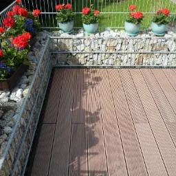 Tarasy z desek kompozytowych, balustrada z siatki gabionowej wypełnionej kamieniami, donice z czerwonymi i niebieskimi kwiatami.