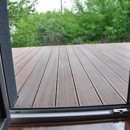 RedWood - Widok z wnętrza przez otwarte drzwi balkonowe na drewniany taras i zielone drzewa w tle.