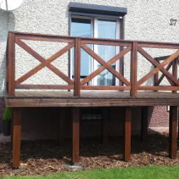 Drewniana antresola z balustradą w stylu rustykalnym, zlokalizowana przy domu w Kalwarii Zebrzydowskiej, z widocznymi elementami wykończenia ogrodu.