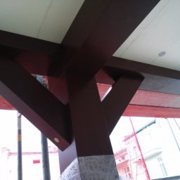 RedWood - Konstrukcja antresoli z brązowych belek drewnianych pod białym stropem, widok z dołu podczas montażu, ochrona folią u podstawy słupa.