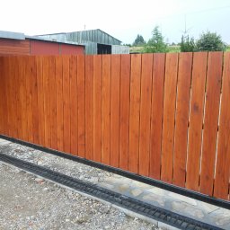 RedWood - Drewniana brama przesuwna z pionowych desek w kolorze brązowym, zamontowana na metalowej szynie z odpływem liniowym, na tle budynków gospodarczych.