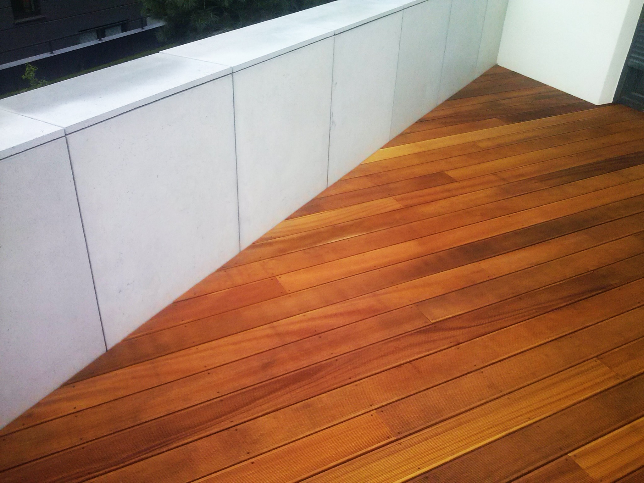 Drewniana podłoga tarasu z desek ułożonych ukośnie, przylegająca do betonowej ściany. Widoczne delikatne usłojenie drewna i szare fugi pomiędzy płytami betonowymi.