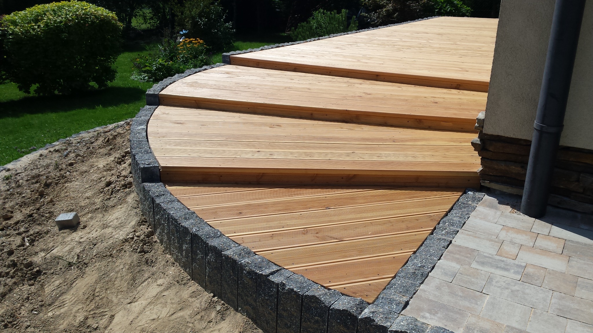 Drewniany taras z trzema stopniami, obrzeżem z szarej kostki i fragmentem elewacji budynku z rynną spustową.