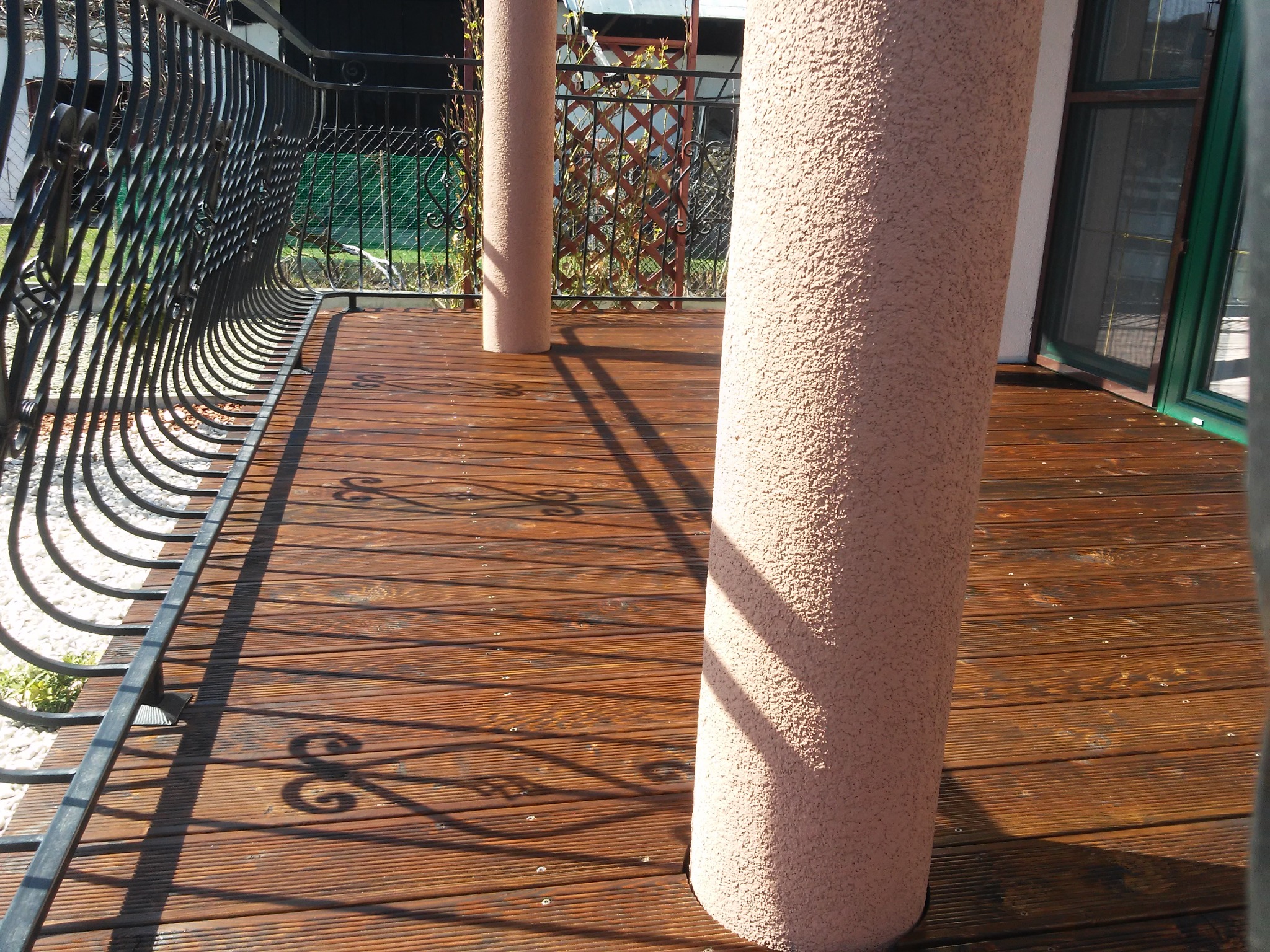 Drewniany taras z ozdobną, kutą balustradą i dwoma okrągłymi, teksturowanymi filarami w kolorze beżowym, oświetlony słonecznym światłem.