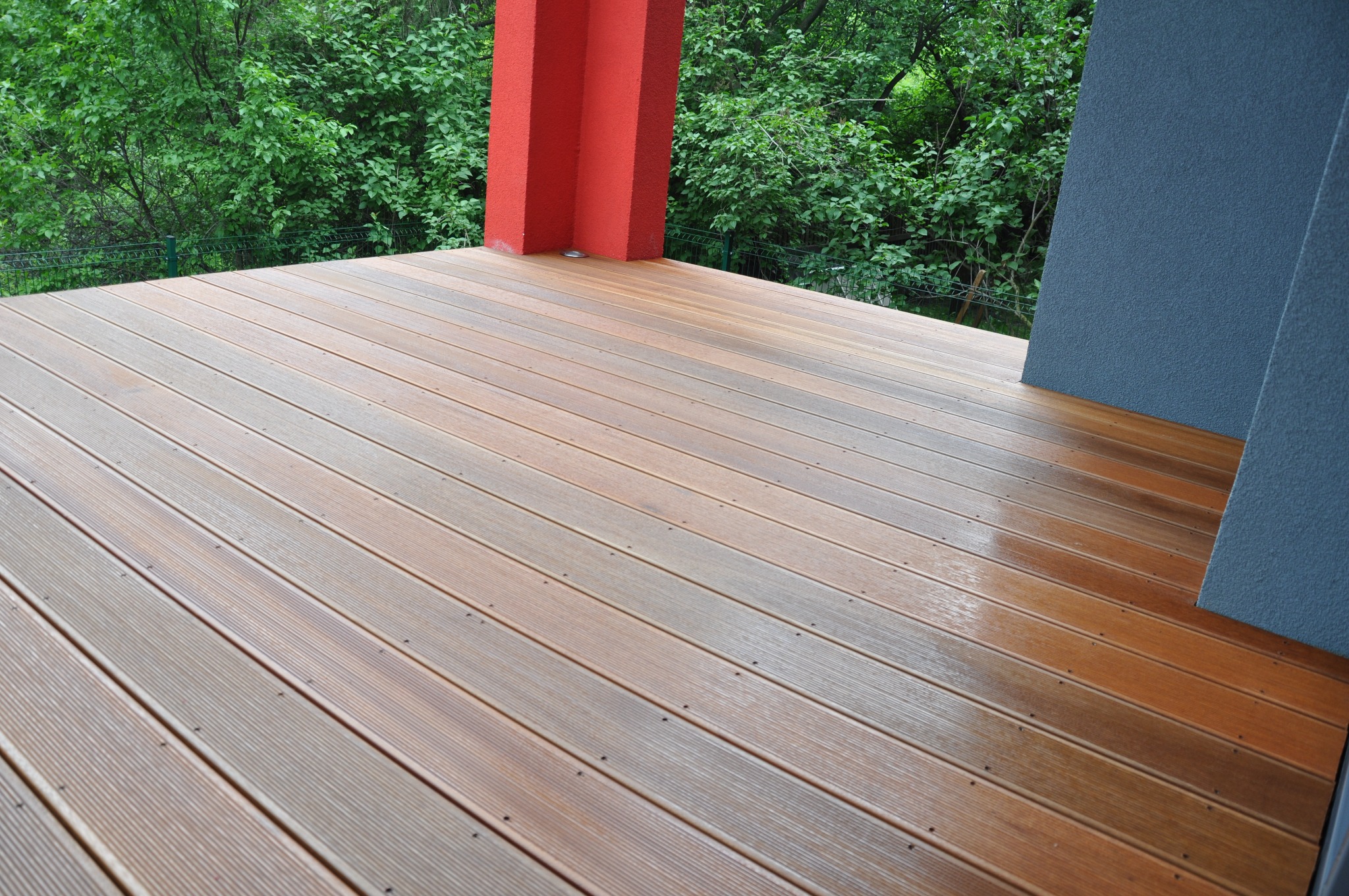 Nowoczesny taras z desek kompozytowych w odcieniach brązu, z widocznym czerwonym słupem i fragmentem szarej ściany budynku, otoczony zielenią drzew.