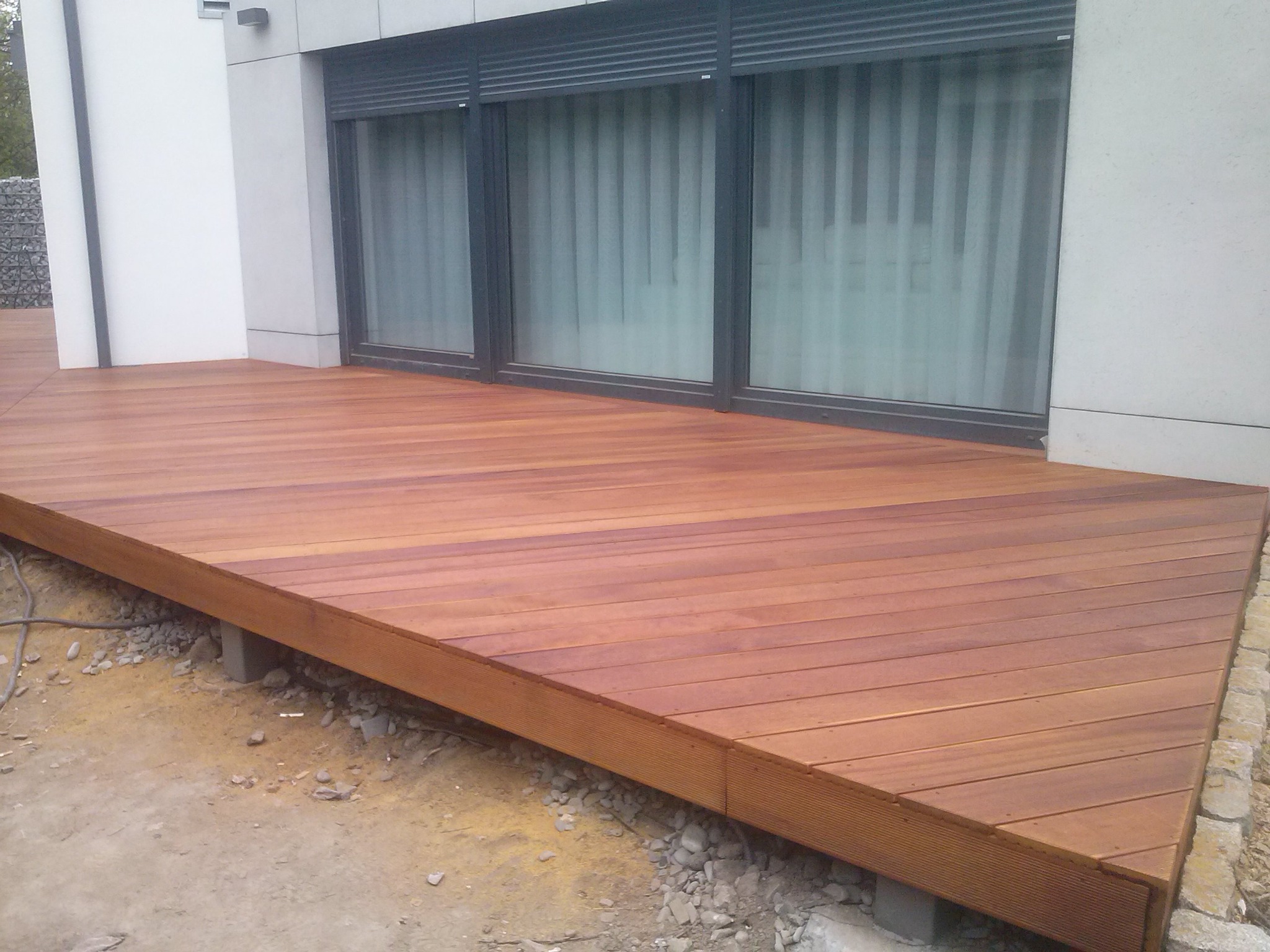 Nowoczesny taras z desek kompozytowych w odcieniach brązu, z widocznymi szarymi oknami z roletami zewnętrznymi i fragmentem białej elewacji budynku w tle, na etapie wykończenia.