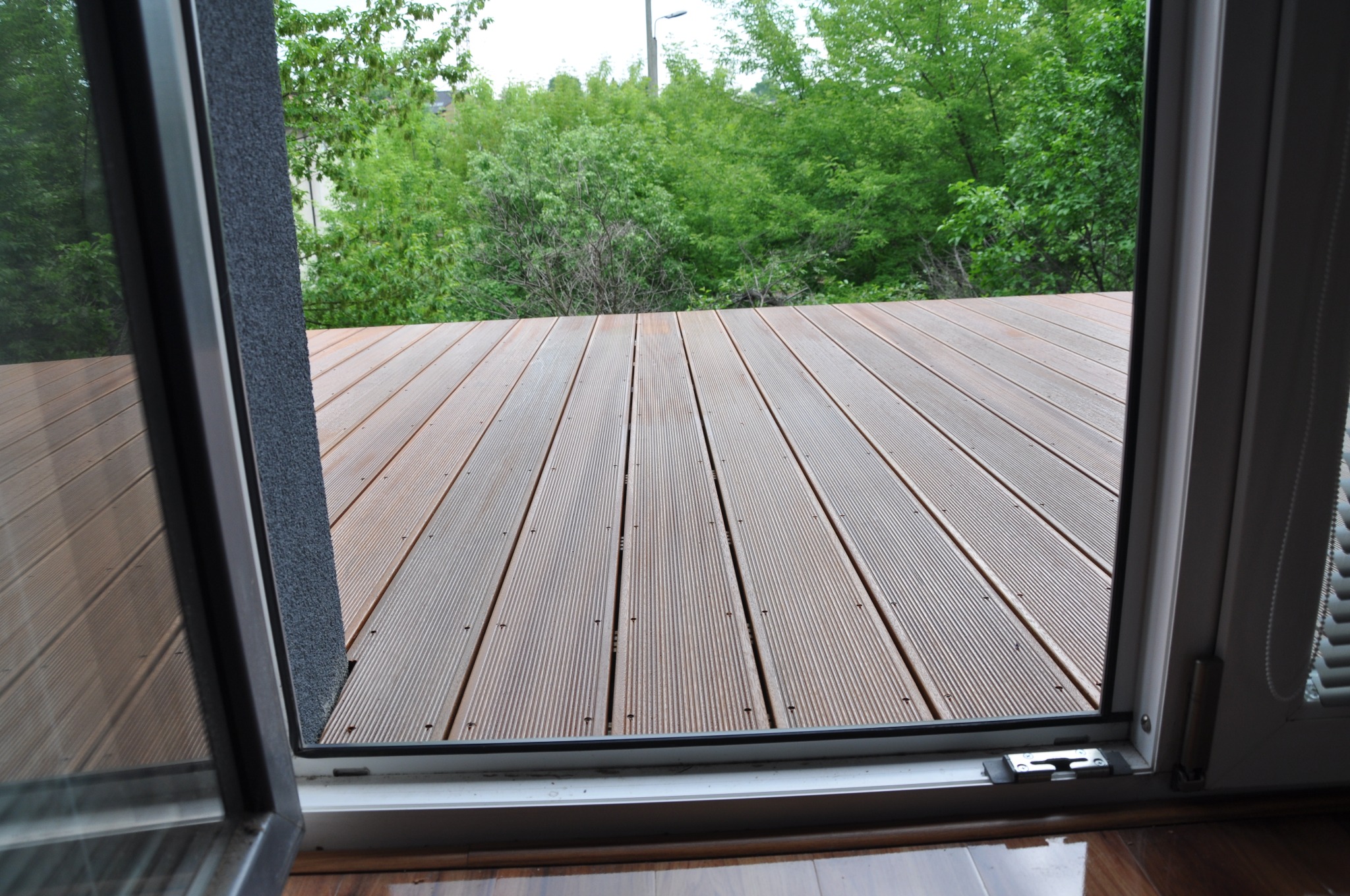 Widok z wnętrza przez otwarte drzwi balkonowe na drewniany taras i zielone drzewa w tle.