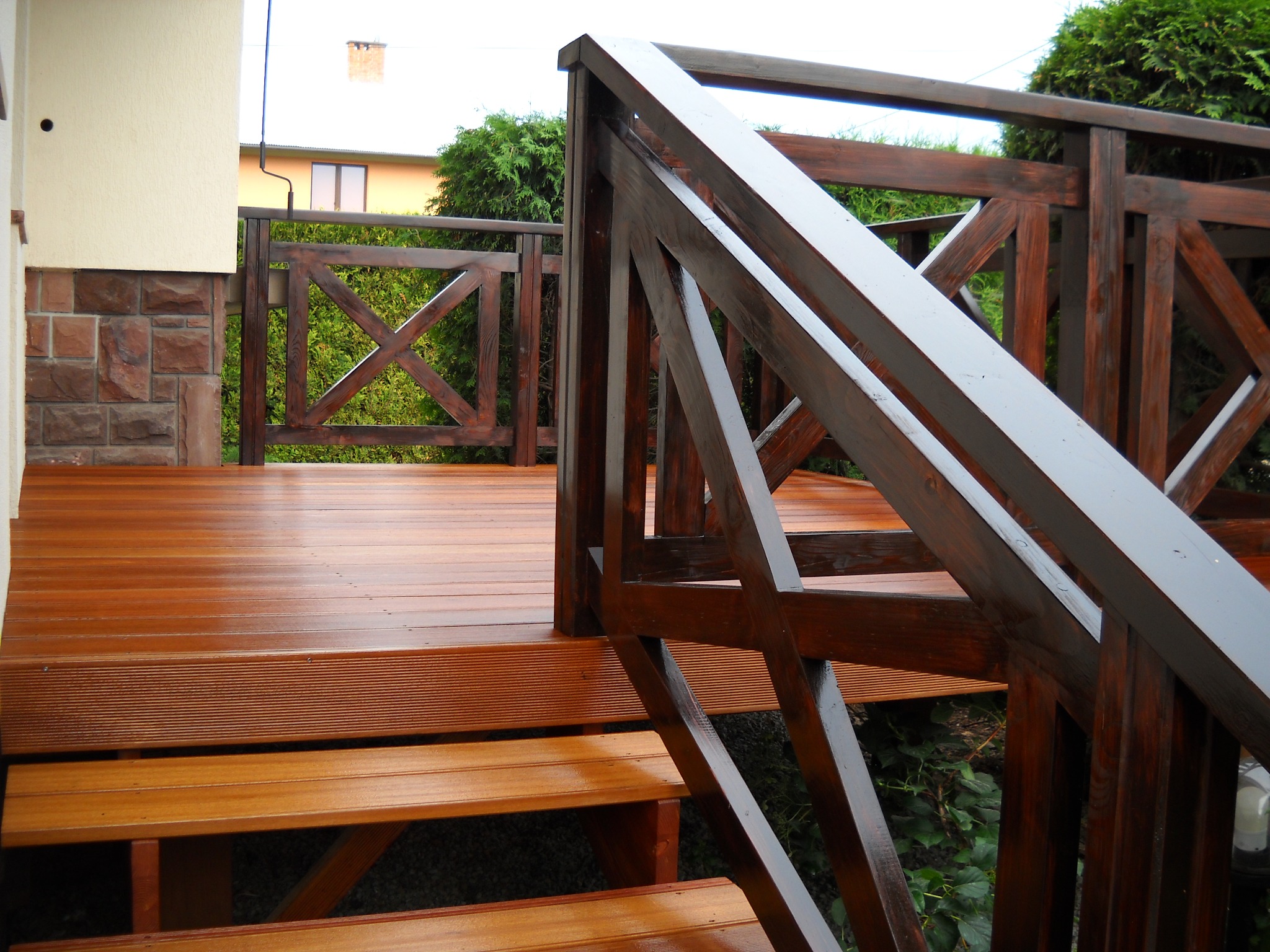 Świeżo polakierowany drewniany taras z poręczami o geometrycznym wzorze krzyża, widok z dołu na schody, z elementami elewacji budynku w tle.