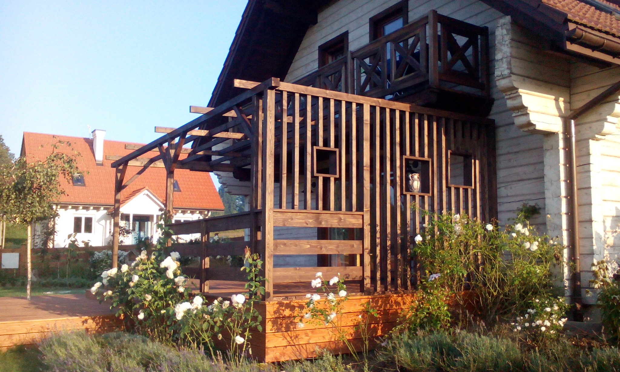 Drewniana weranda z ażurową konstrukcją i wstawionymi kwadratowymi ramkami, przylegająca do domu z drewnianą elewacją, z widocznym balkonem i donicą w jednej z ramek, otoczona kwitnącymi różami...