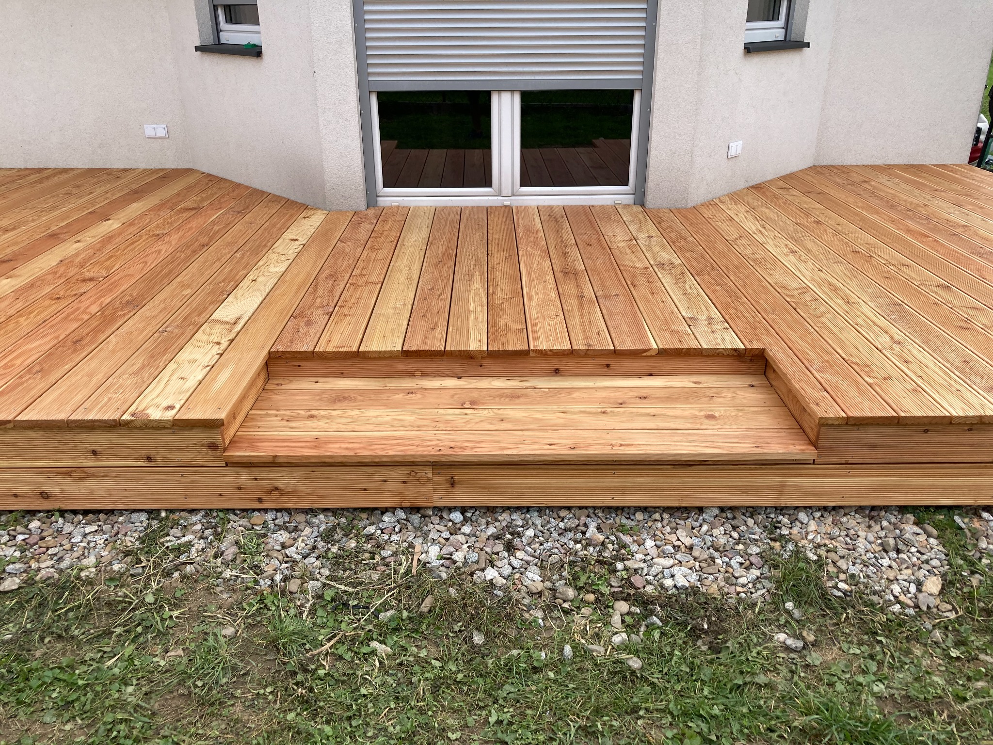 Nowoczesny taras z desek kompozytowych w odcieniach ciepłego brązu, z niskimi schodami prowadzącymi do domu z zamkniętymi roletami i jasną elewacją. Pod tarasem widoczne kamienie i trawa.