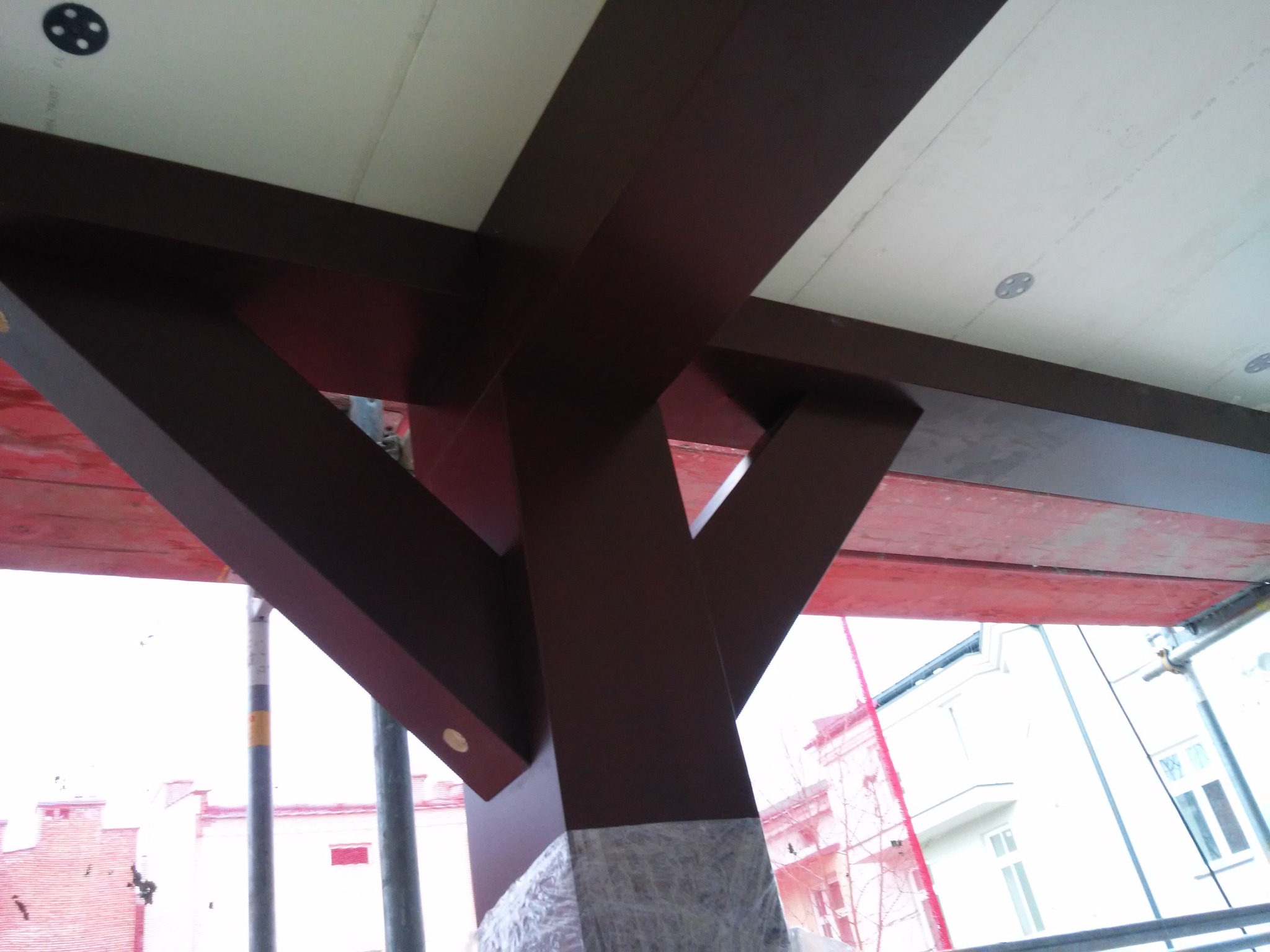 Konstrukcja antresoli z brązowych belek drewnianych pod białym stropem, widok z dołu podczas montażu, ochrona folią u podstawy słupa.