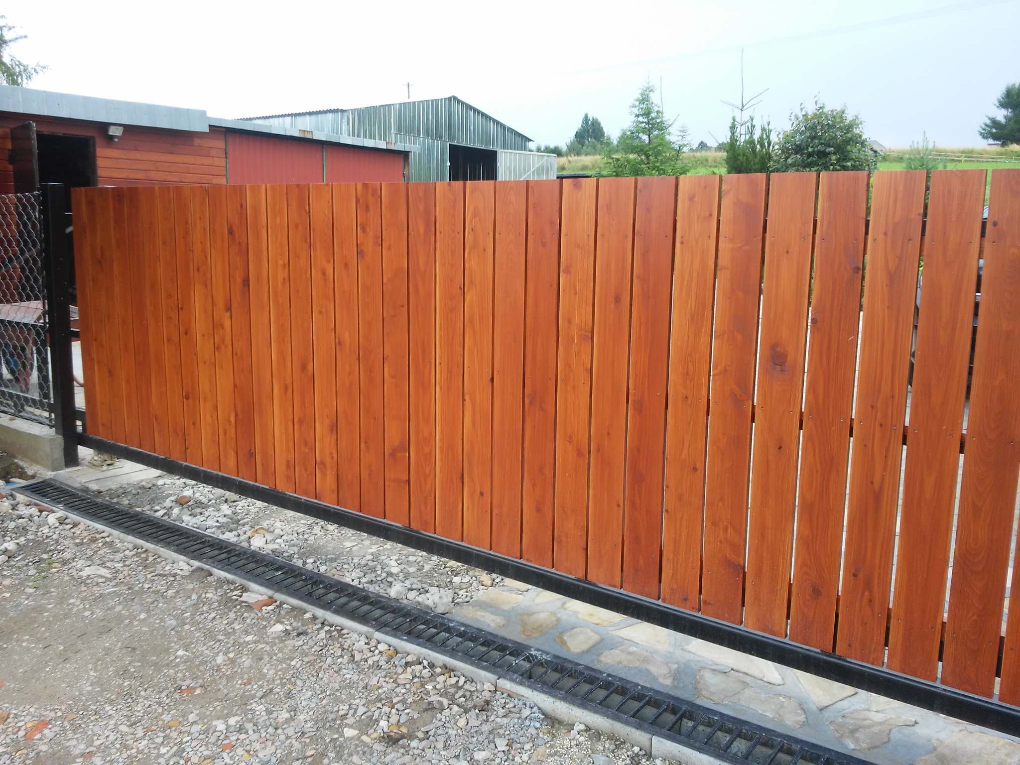 Drewniana brama przesuwna z pionowych desek w kolorze brązowym, zamontowana na metalowej szynie z odpływem liniowym, na tle budynków gospodarczych.