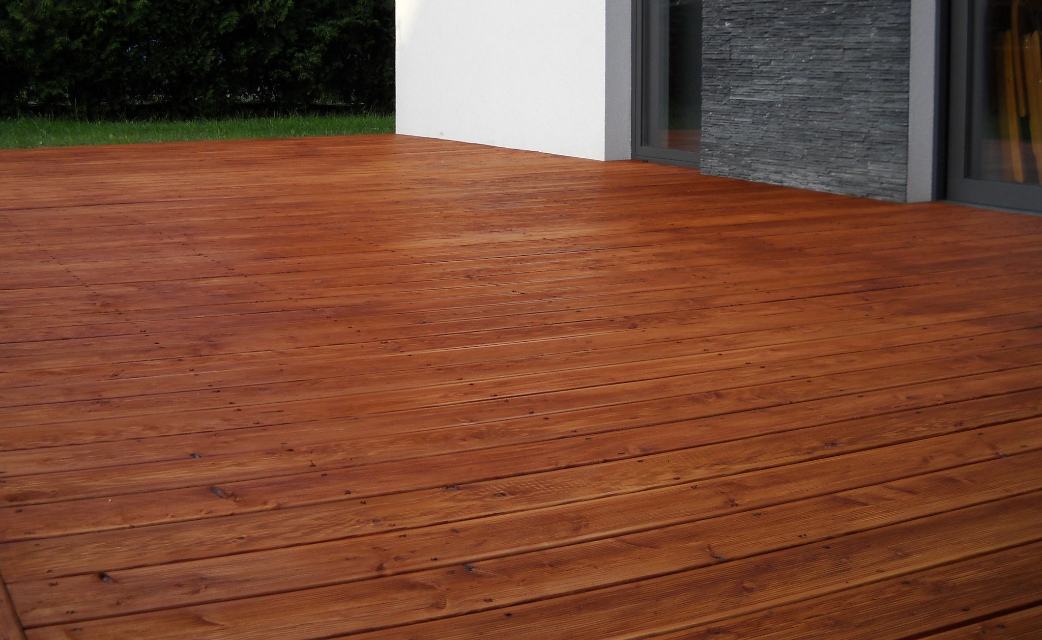 Nowo zaimpregnowany, drewniany taras z deskami o ciepłym, brązowym odcieniu, przylegający do nowoczesnego domu z białymi ścianami i elementami z szarego kamienia.