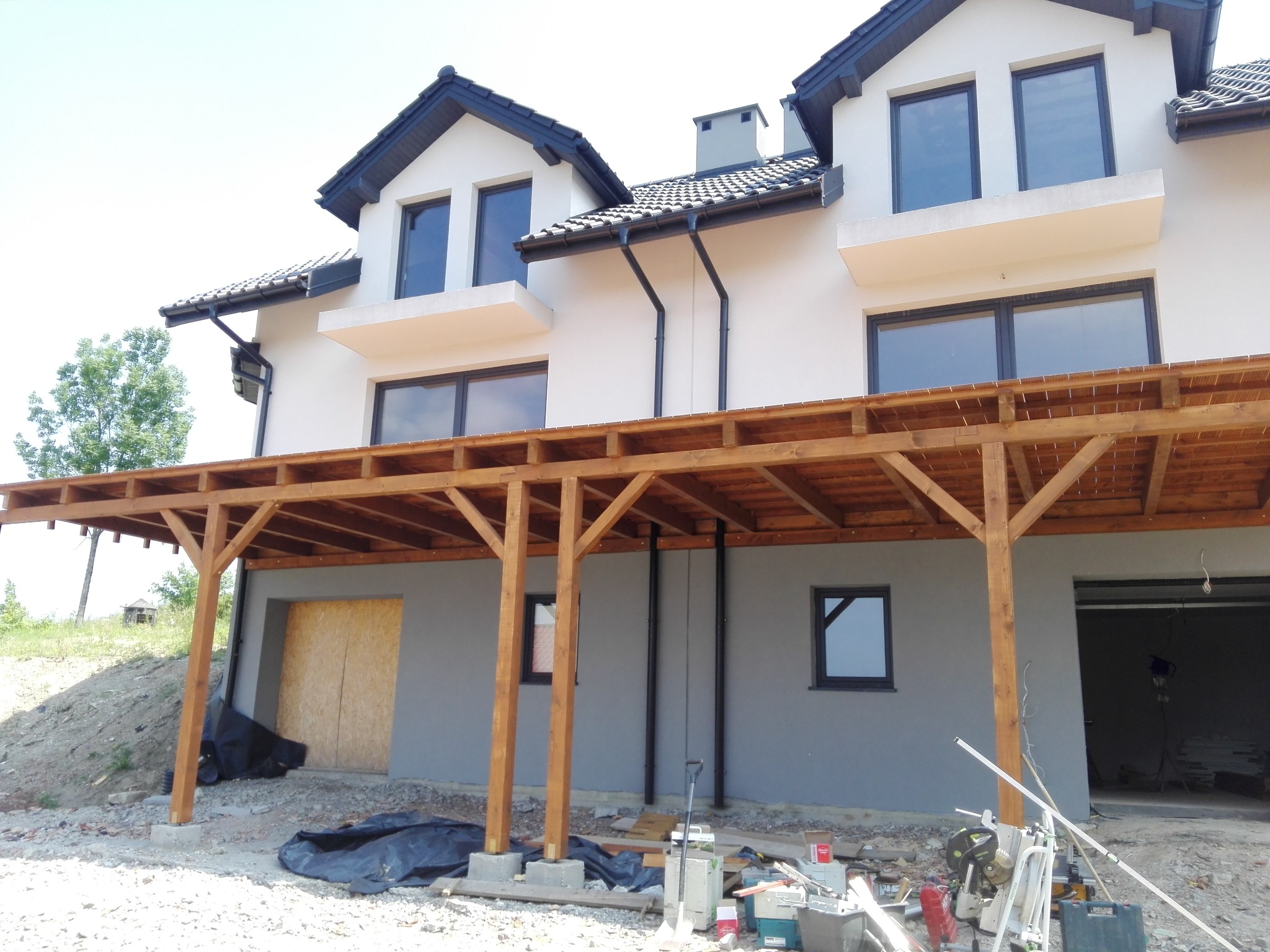 Surowa drewniana konstrukcja zadaszenia tarasu przy nowym, dwukondygnacyjnym domu z szarą elewacją, widoczne narzędzia i materiały budowlane na ziemi.