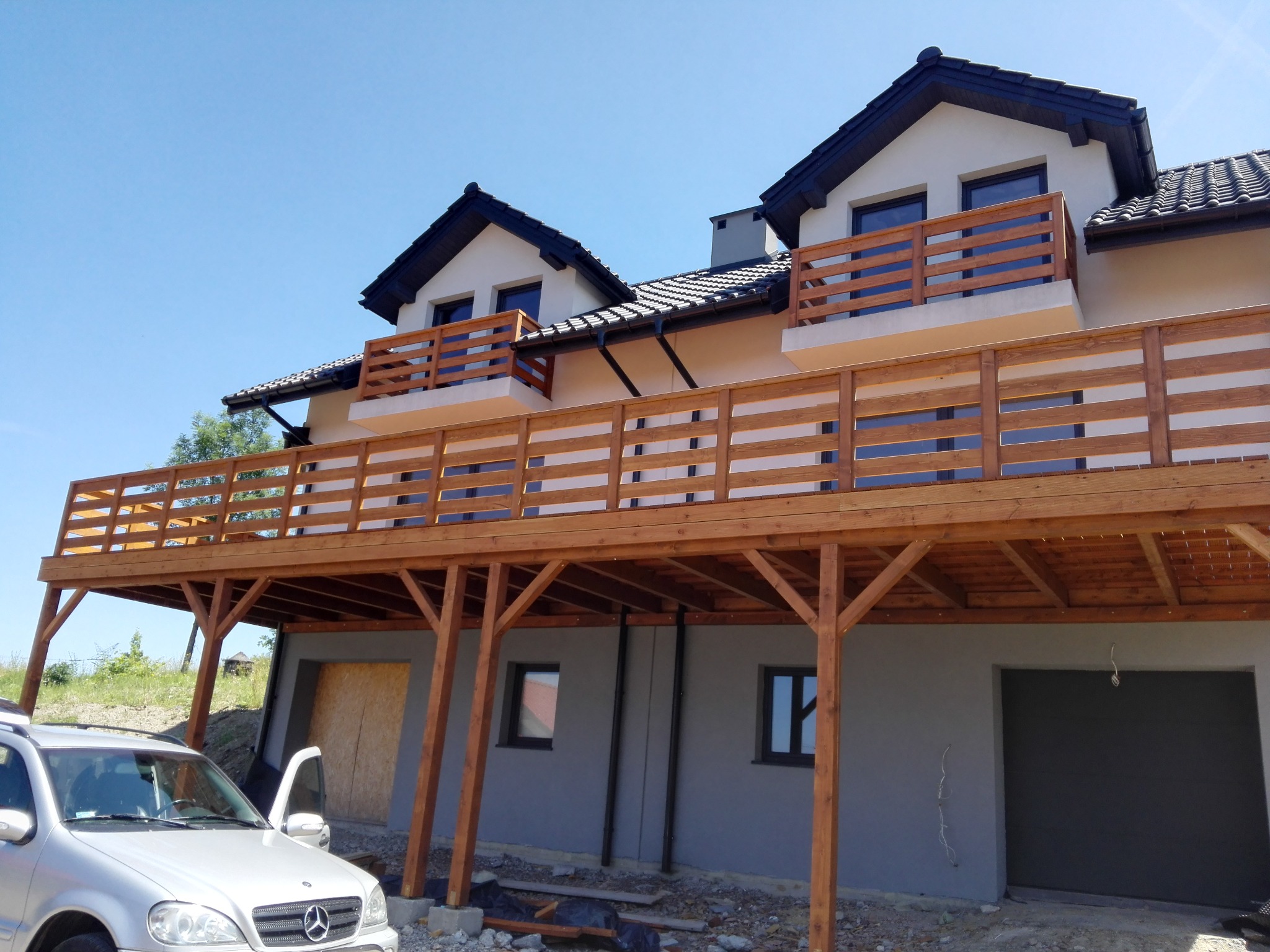 Dwukondygnacyjny dom z drewnianym tarasem i balkonami, widok z dołu, z zaparkowanym samochodem osobowym na pierwszym planie.