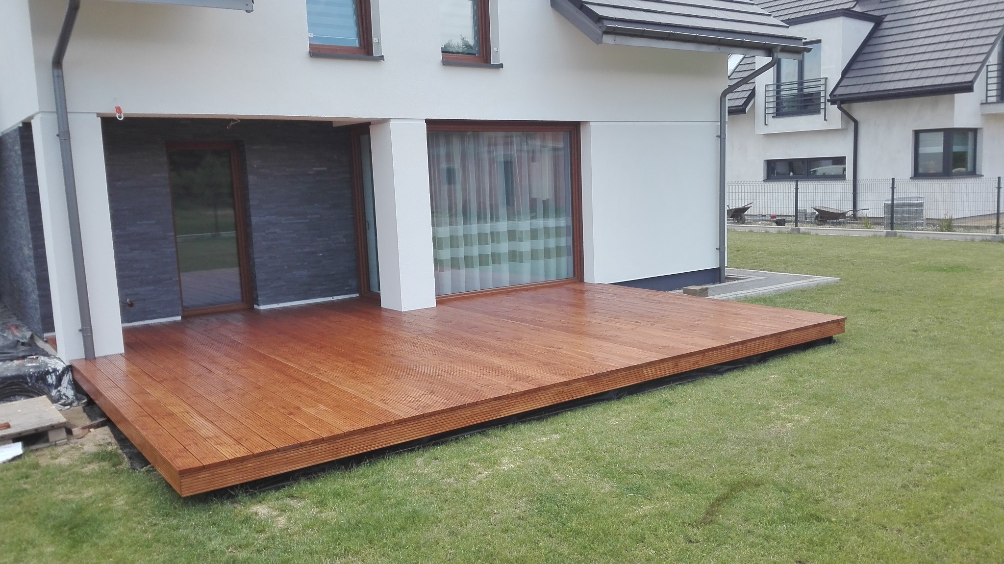 Nowoczesny taras z drewna egzotycznego przylegający do białego domu z szarymi elementami wykończeniowymi, widok na zadbany trawnik i sąsiedni budynek w tle.