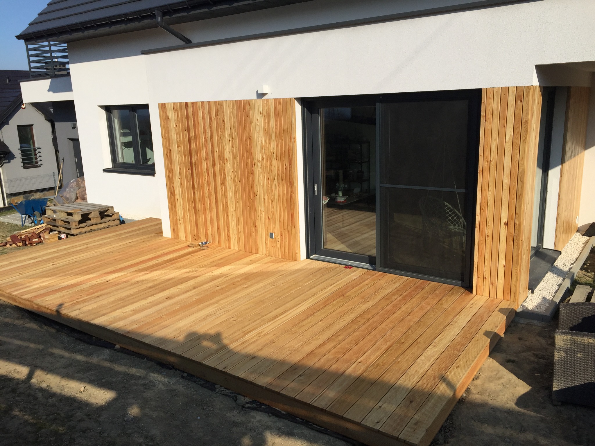 Nowoczesny taras z drewna, z widoczną deską elewacyjną z tego samego materiału, przylegający do białego budynku z dużym przesuwnym oknem.