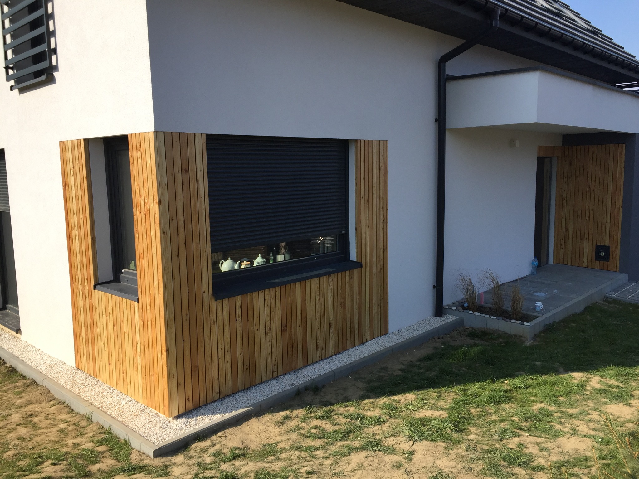 Nowoczesny dom z drewnianą elewacją okienną i drzwiami wejściowymi, obłożony jasnym drewnem, z roletami zewnętrznymi i kamiennym obrzeżem.