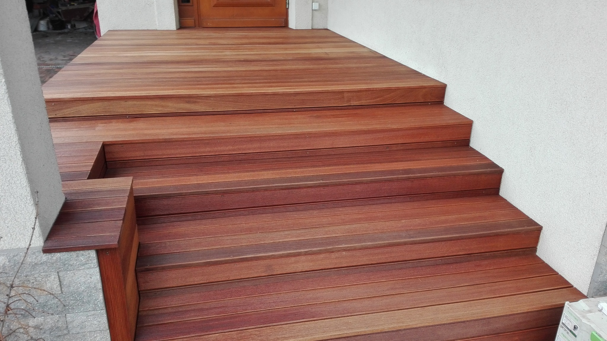 Drewniany taras z ryflowanych desek prowadzący do drzwi wejściowych, z niską barierką po lewej stronie, przylegający do ściany budynku o szorstkiej fakturze.