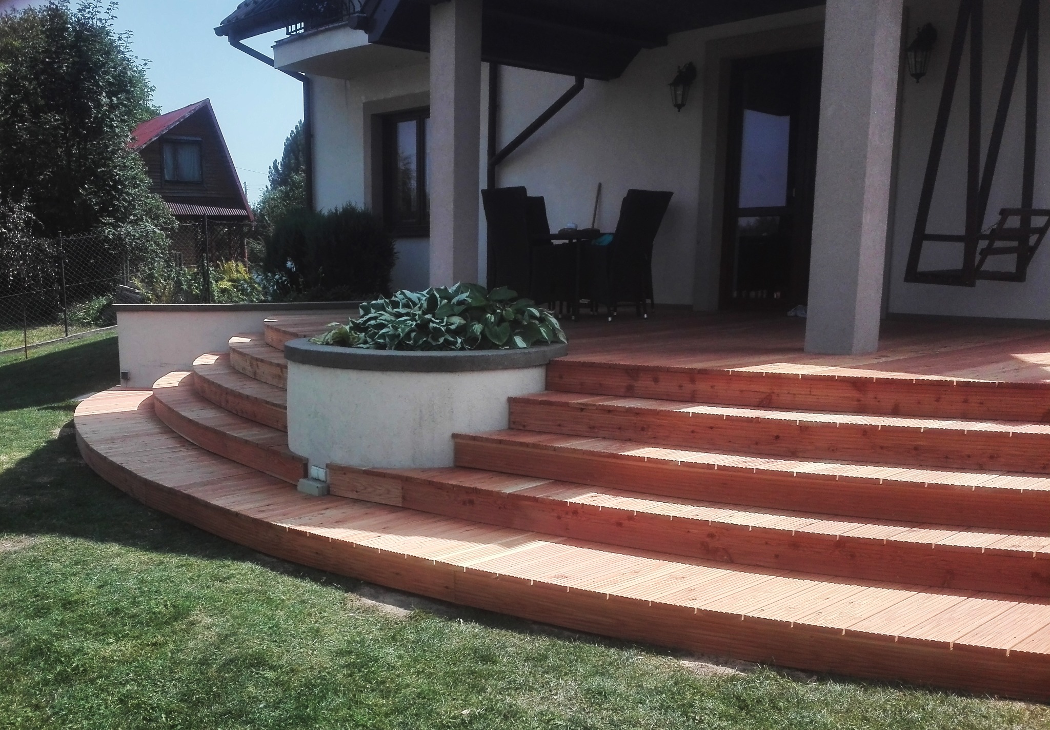 Drewniane schody tarasowe prowadzące do domu, z okrągłą donicą z roślinami po lewej stronie, trawnik przed schodami, dom w tle.