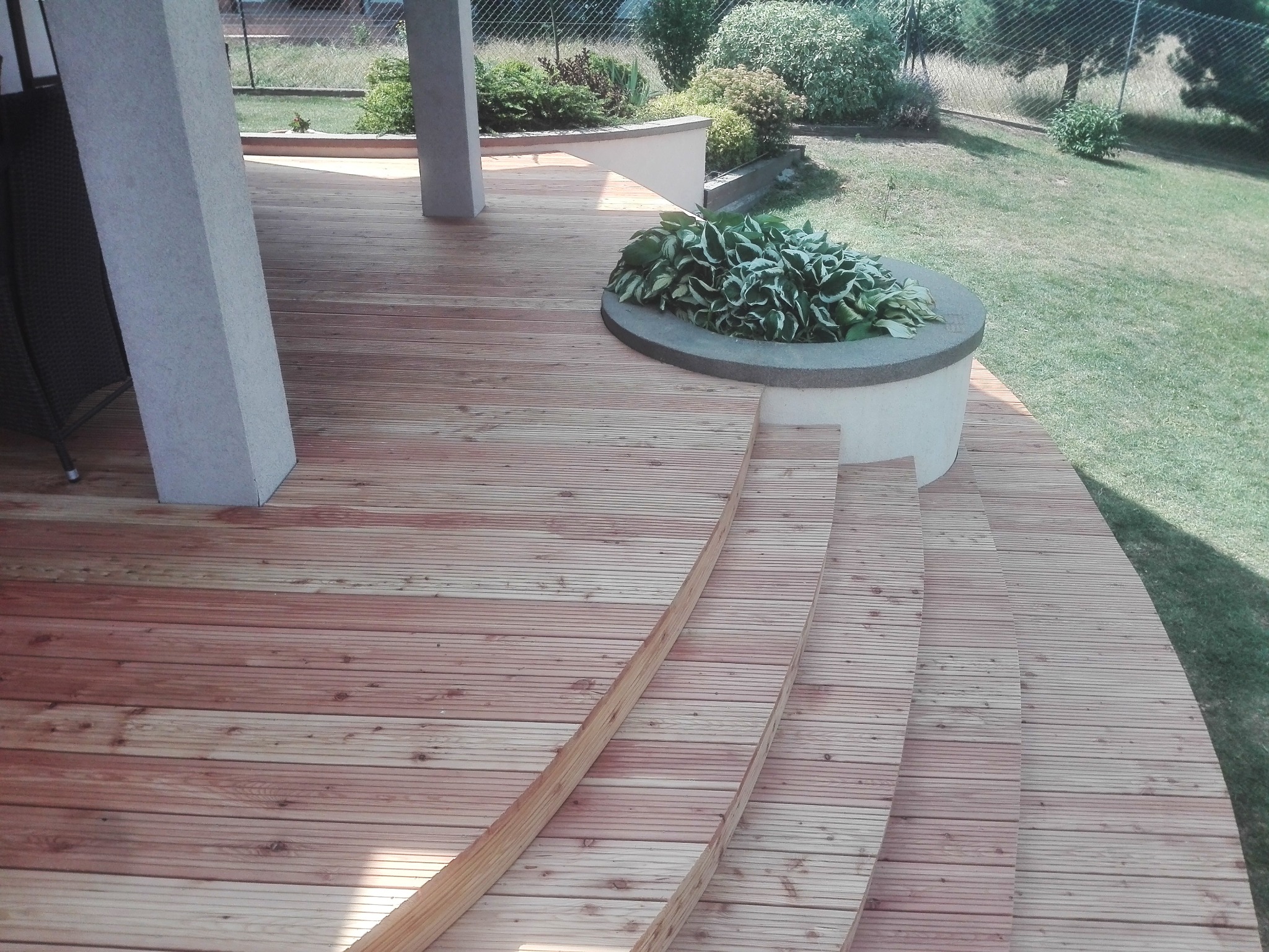 Drewniany taras z desek z zaokrąglonymi schodami i okrągłą donicą z roślinami, widoczne dwie betonowe podpory i trawnik w tle.