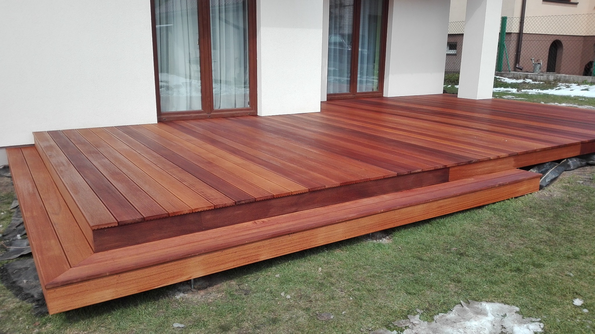 Nowoczesny taras z egzotycznego drewna z dwoma stopniami, przylegający do białego budynku z brązowymi ramami okiennymi, na trawniku z resztkami śniegu.