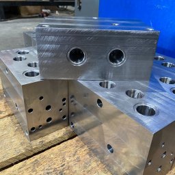 TOSE Tomasz Stach - Precyzyjnie obrobione, metalowe bloki z licznymi otworami i gwintami, ułożone na drewnianej powierzchni. Widoczne ślady obróbki skrawaniem na powierzchni bloków.