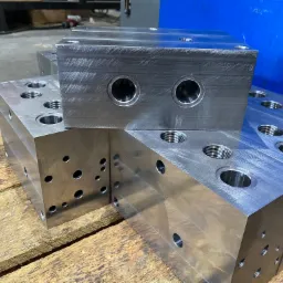 Precyzyjnie obrobione, metalowe bloki z licznymi otworami i gwintami, ułożone na drewnianej powierzchni. Widoczne ślady obróbki skrawaniem na powierzchni bloków.