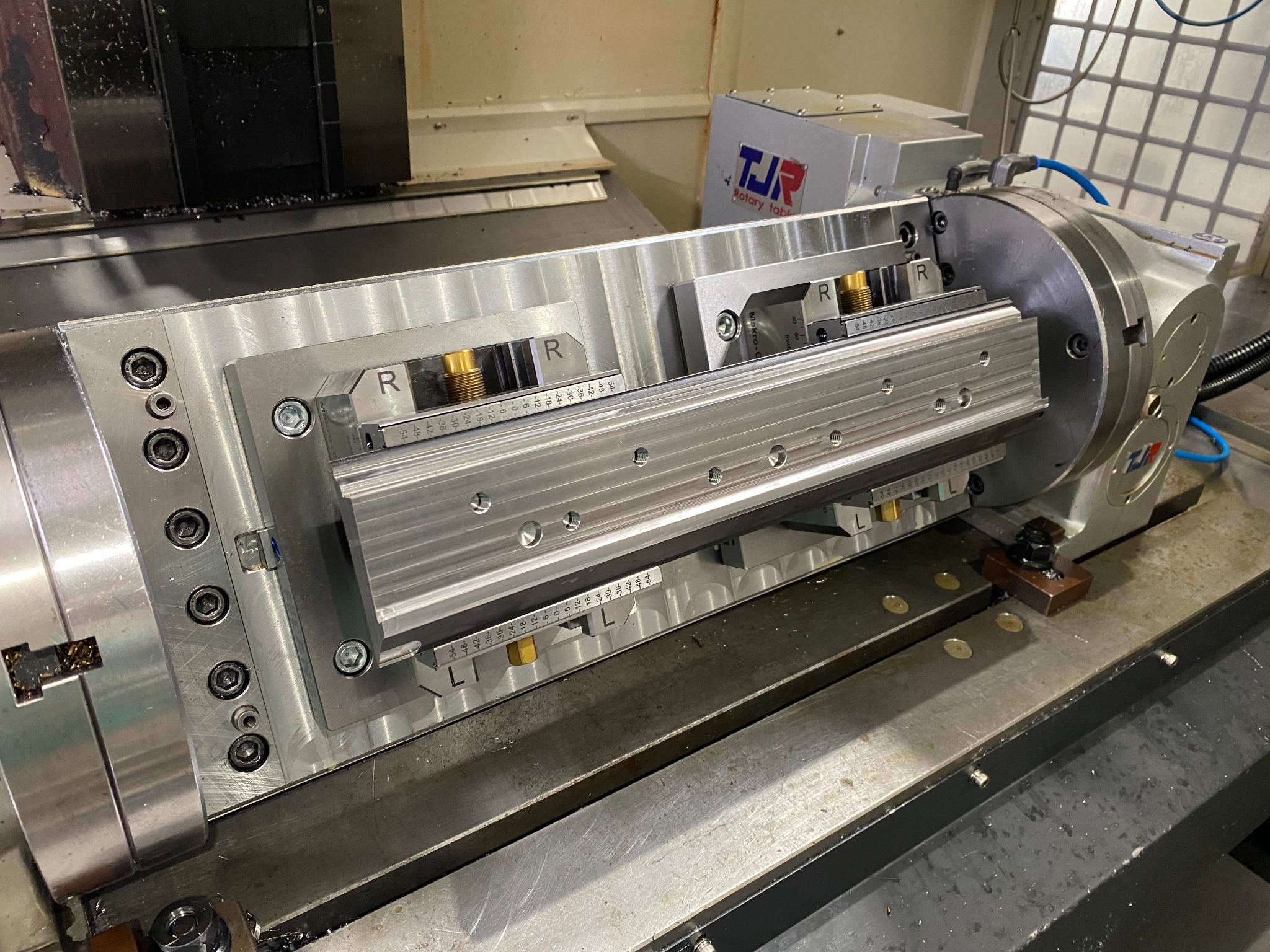 Szczegółowe ujęcie frezarki CNC z zamocowanym obrotowym stołem i uchwytem, widoczne oznaczenia 'R' i 'L' na elementach mocujących, oraz logo 'TJR Rotary table' w tle.