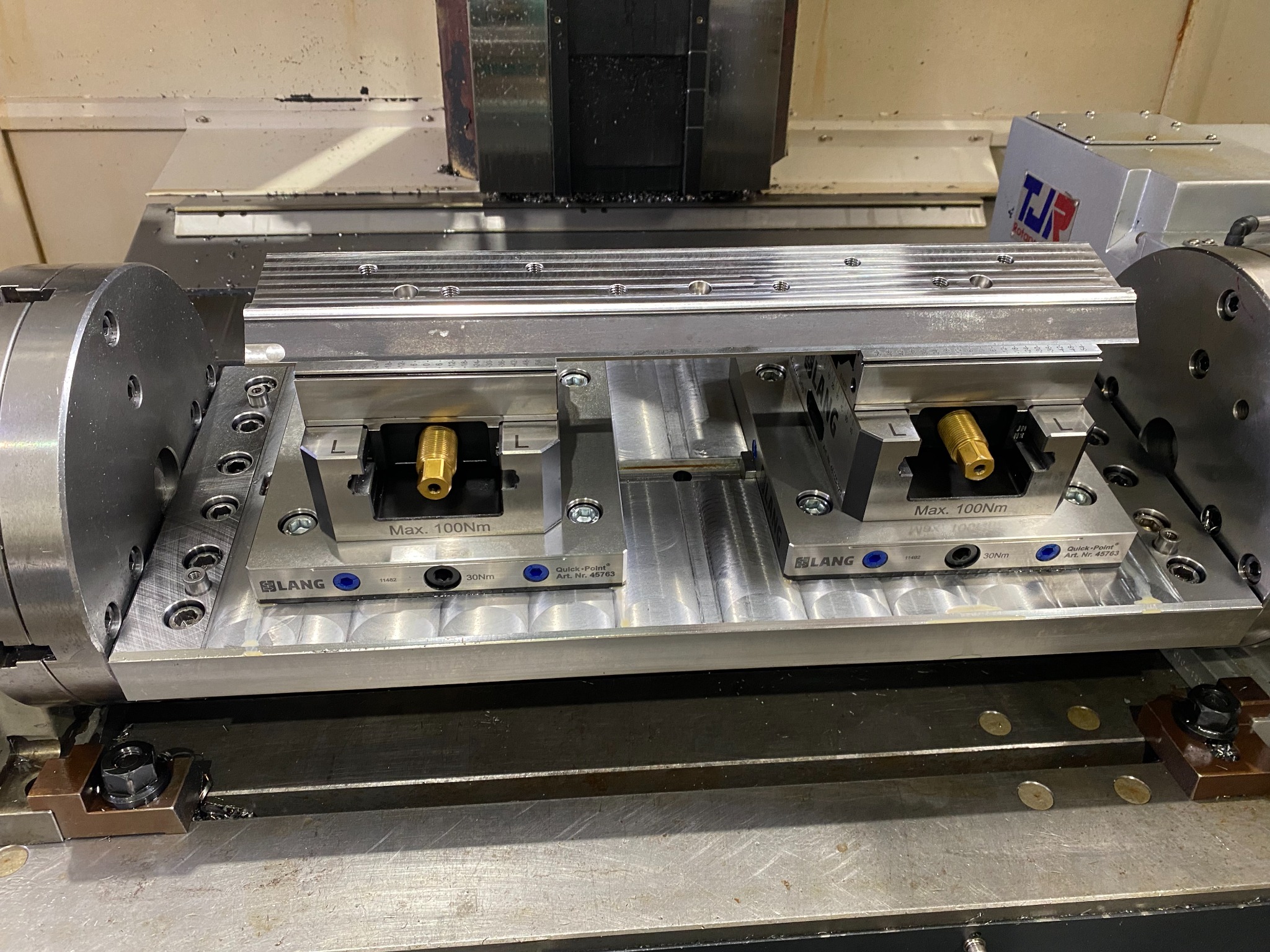 Obróbka CNC: Detal zamocowany w precyzyjnym uchwycie LANG Quick-Point na stole frezarki CNC, widoczne wióry po obróbce.