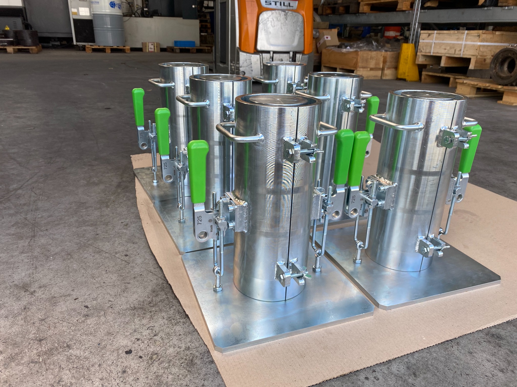Seryjnie wykonane metalowe cylindryczne formy z zielonymi dźwigniami mocującymi, ustawione na metalowych podstawach, widoczne w hali produkcyjnej z wózkiem widłowym w tle.