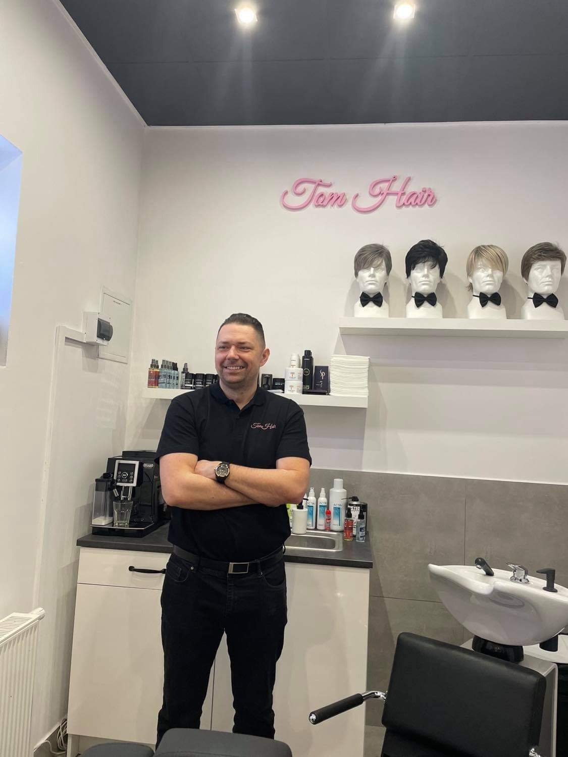 Uśmiechnięty mężczyzna w czarnej koszulce z logo firmy, stojący na tle wnętrza salonu z umywalką fryzjerską, ekspres do kawy i czterema manekinami z perukami na półce pod neonem 'Tom Hair'.