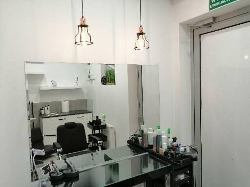 Wnętrze salonu z stanowiskiem pracy, widoczne lustro, fotel fryzjerski, w tle szafki i akcesoria, oświetlenie w stylu industrialnym.