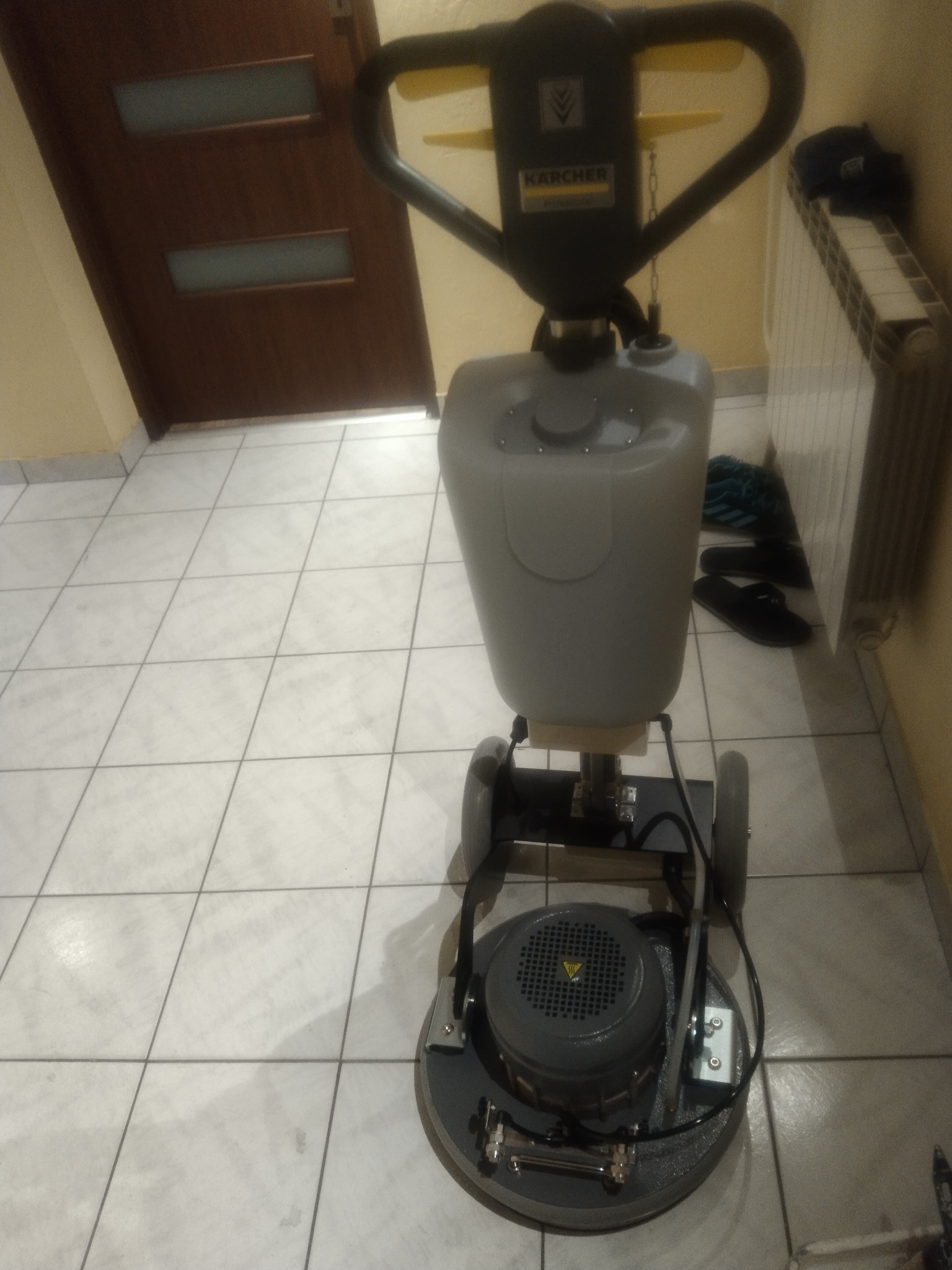 Profesjonalna maszyna Karcher do czyszczenia podłóg na białych płytkach. Widoczny zbiornik na wodę i uchwyt. Obok drzwi i grzejnik.