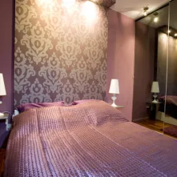 Sypialnia w odcieniach fioletu z wzorzystą tapetą za łóżkiem, lampkami nocnymi na półkach i szafą z lustrzanymi drzwiami.