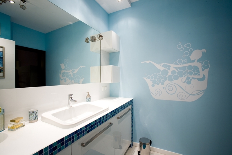 Jasna łazienka z błękitnymi ścianami, białą umywalką wpuszczoną w blat wykończony mozaiką, lustrem i naklejką ścienną przedstawiającą kobietę w wannie pełnej piany.