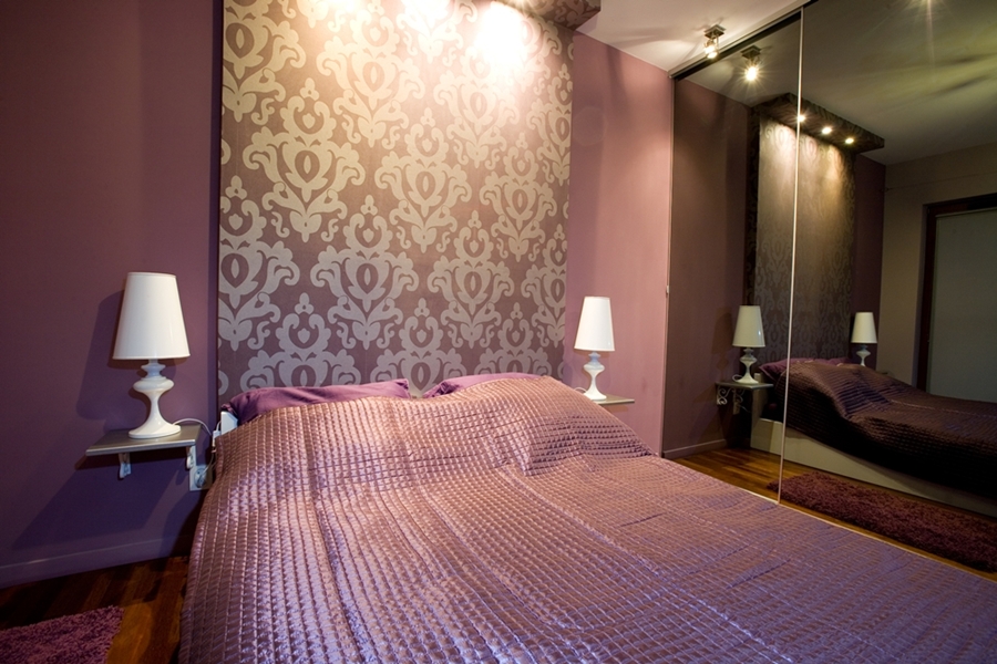 Sypialnia w odcieniach fioletu z wzorzystą tapetą za łóżkiem, lampkami nocnymi na półkach i szafą z lustrzanymi drzwiami.