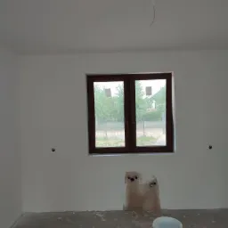 Pokój w trakcie remontu: świeżo pomalowane ściany na biało, widoczne puszki instalacyjne, niezabezpieczona podłoga i wiaderka z farbą.