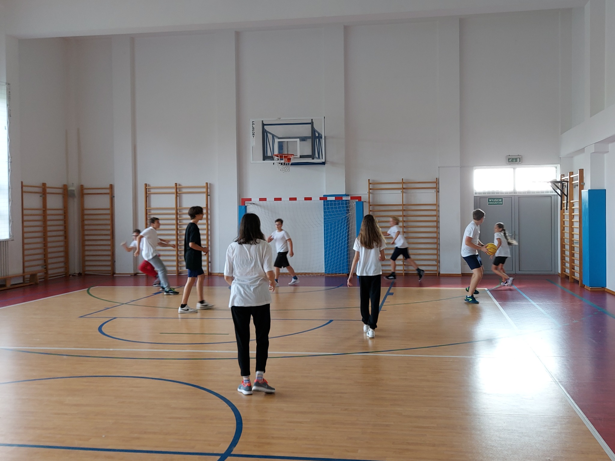 Grupa nastolatków ćwiczy w hali sportowej z parkietem i koszem do koszykówki, widoczne drabinki gimnastyczne przy ścianie.
